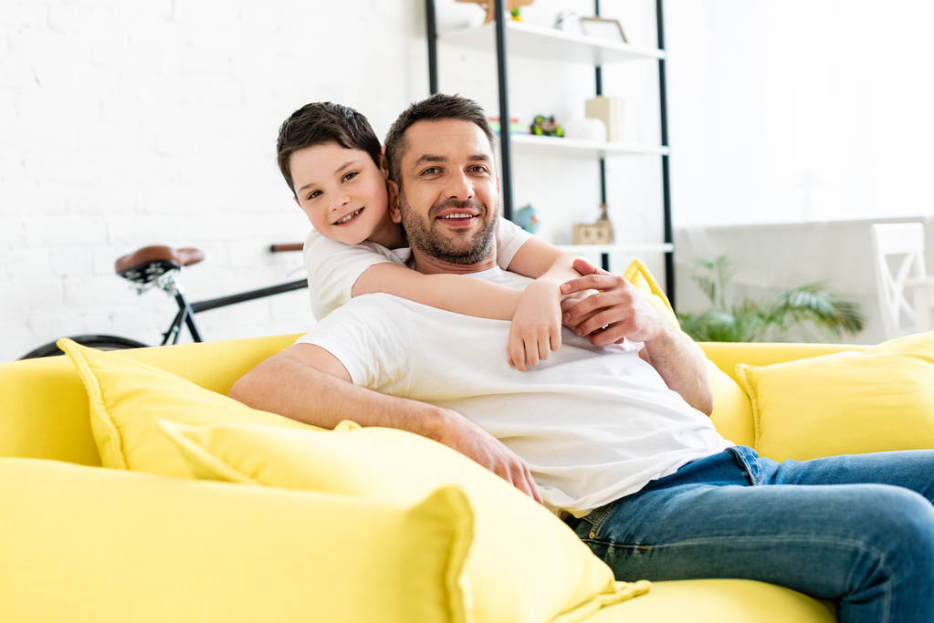 son looking at camera while hugging father sitting on couch at home - Photo, Image