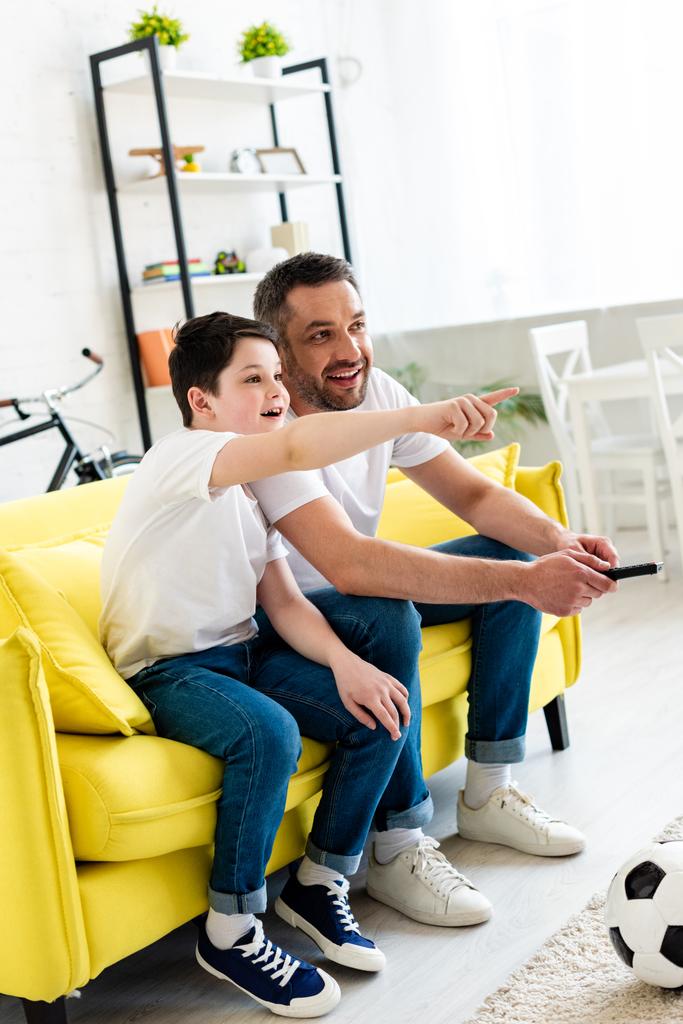 Sohn zeigt mit dem Finger, während er auf Couch sitzt und mit Vater zu Hause fernsieht - Foto, Bild