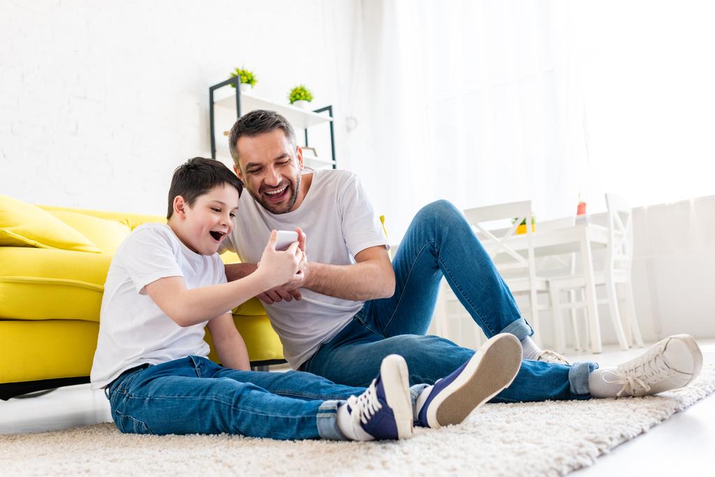 podekscytowany ojciec i syn siedzi na dywanie i za pomocą smartfona w salonie - Zdjęcie, obraz