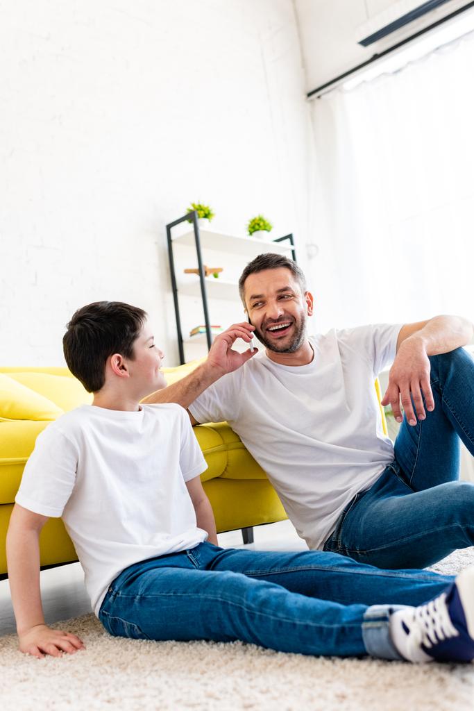 Syn siedzi na dywanie w pobliżu ojca rozmawiając na smartfonie w salonie - Zdjęcie, obraz