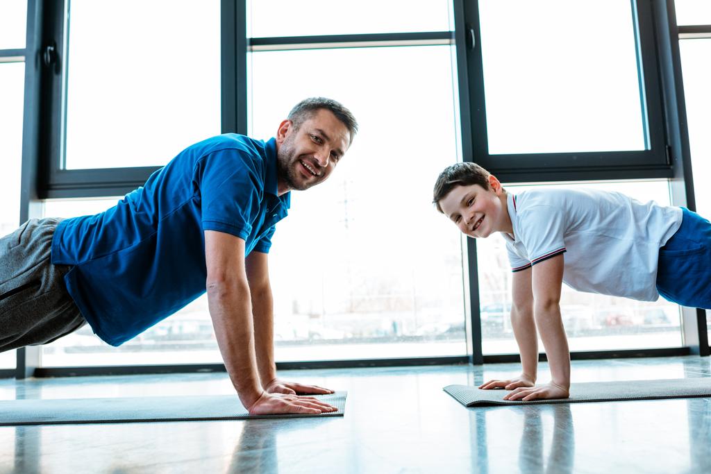 happy father and son looking at camera while doing push up exercise at gym - Photo, Image
