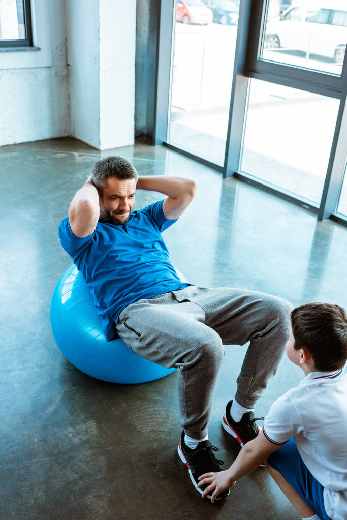 son helping father sitting on fitness ball and doing sit up exercise at gym - Photo, Image
