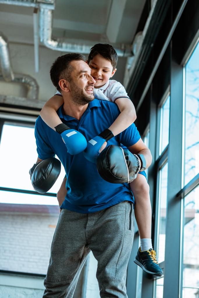 feliz padre dando paseo a cuestas al hijo sonriente en guantes de boxeo en el gimnasio
 - Foto, imagen