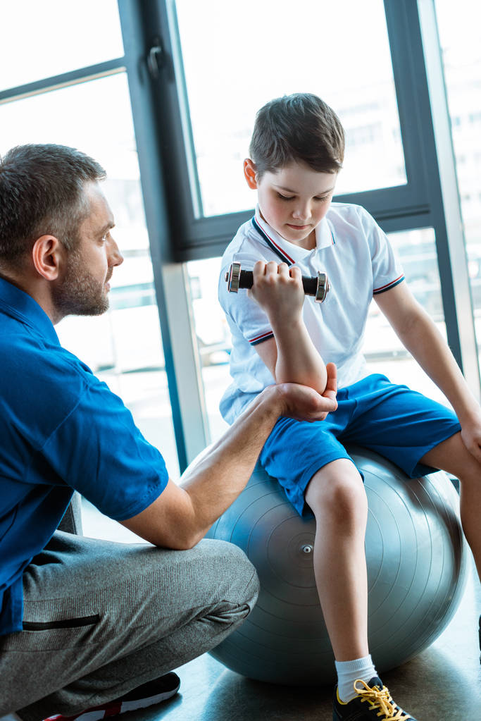 vader helpt zoon op fitness mat oefenen met halter in sportcentrum - Foto, afbeelding