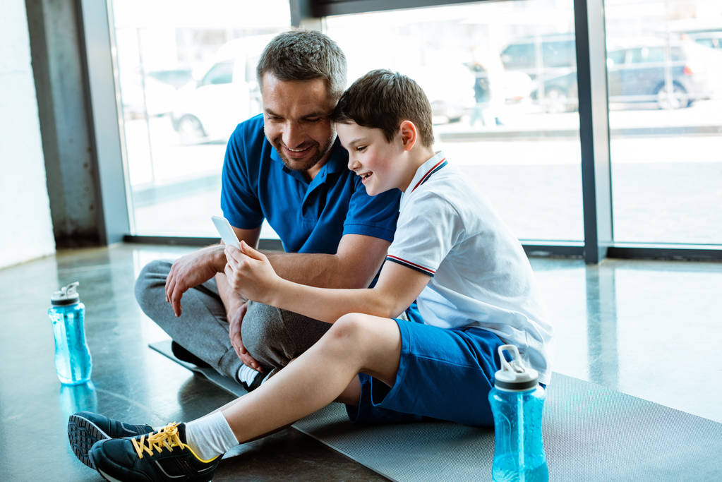 apa és fia ül a fitness mat és használata okostelefon a tornateremben - Fotó, kép