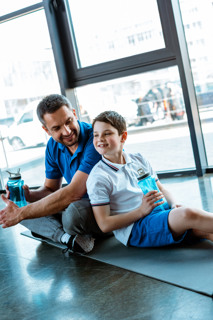 heureux père et fils assis sur tapis de fitness avec des bouteilles de sport à la salle de gym
 - Photo, image