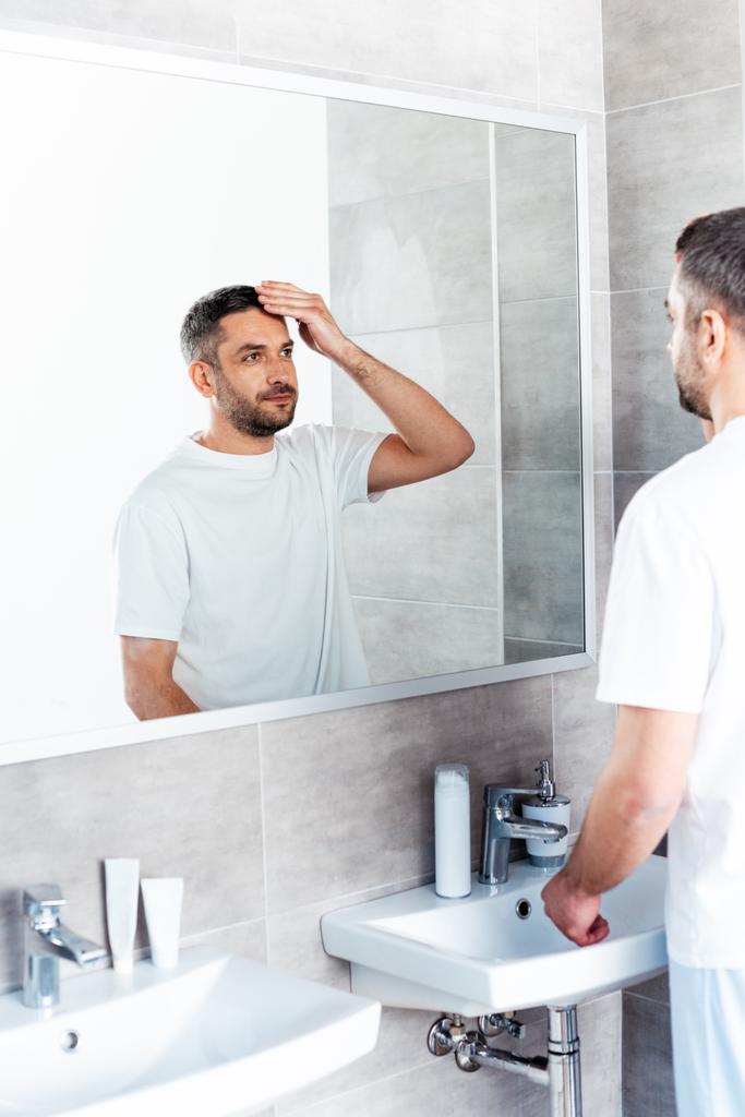 przystojny mężczyzna patrząc na lustro i dostosowanie fryzury w łazience - Zdjęcie, obraz