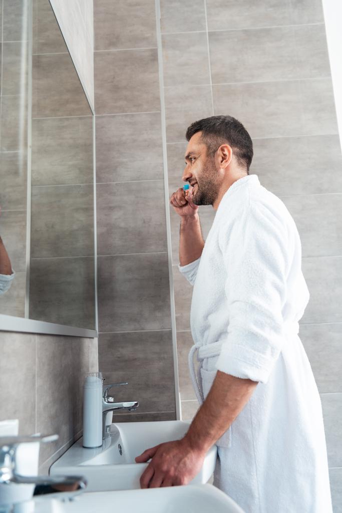 hombre guapo de albornoz blanco cepillarse los dientes en el baño durante la rutina de la mañana
 - Foto, Imagen