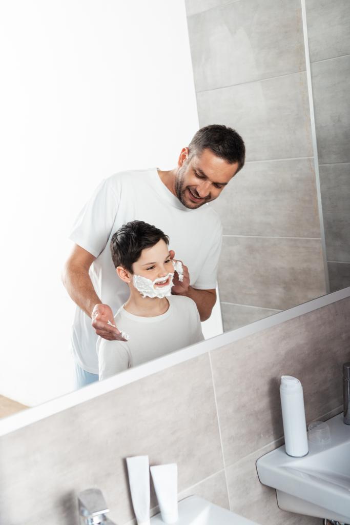 Lächelnder Vater trägt Rasierschaum auf Gesicht seines Sohnes im Badezimmer auf - Foto, Bild