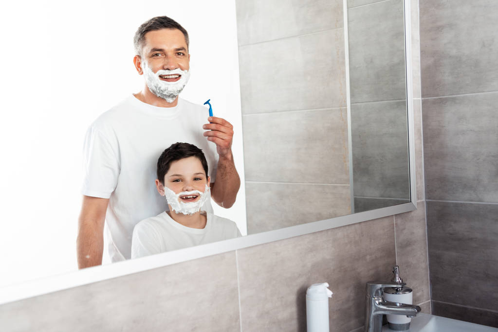 lachende zoon en vader met scheercrème op gezichten kijken naar camera in badkamer - Foto, afbeelding