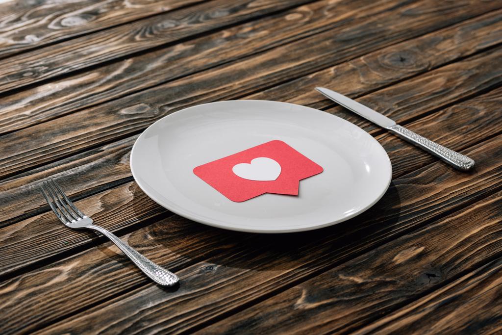 rood papier knippen kaart met hart symbool op witte plaat in de buurt van mes en vork op bruin houten oppervlak - Foto, afbeelding