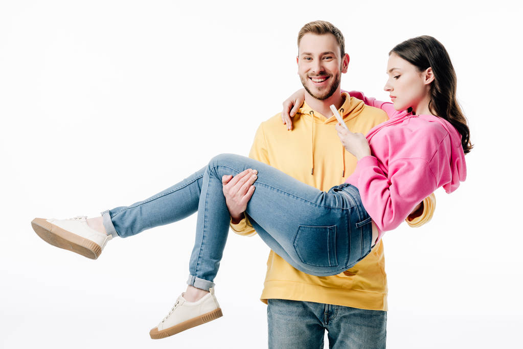 smiling boyfriend looking at camera while holding girl using smartphone isolated on white - Photo, Image