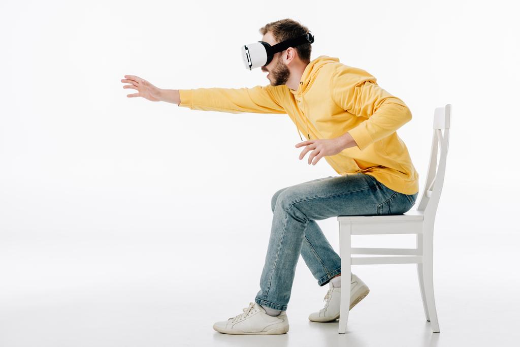 jonge man in virtual reality headset zitten op stoel met uitgestrekte hand op witte achtergrond - Foto, afbeelding