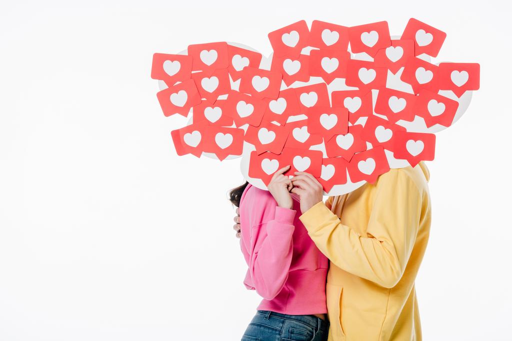 mladý muž a žena v jasných mikinách skrývající se za tvářemi za červeným papírem řezané karty se symboly srdce izolované na bílém - Fotografie, Obrázek