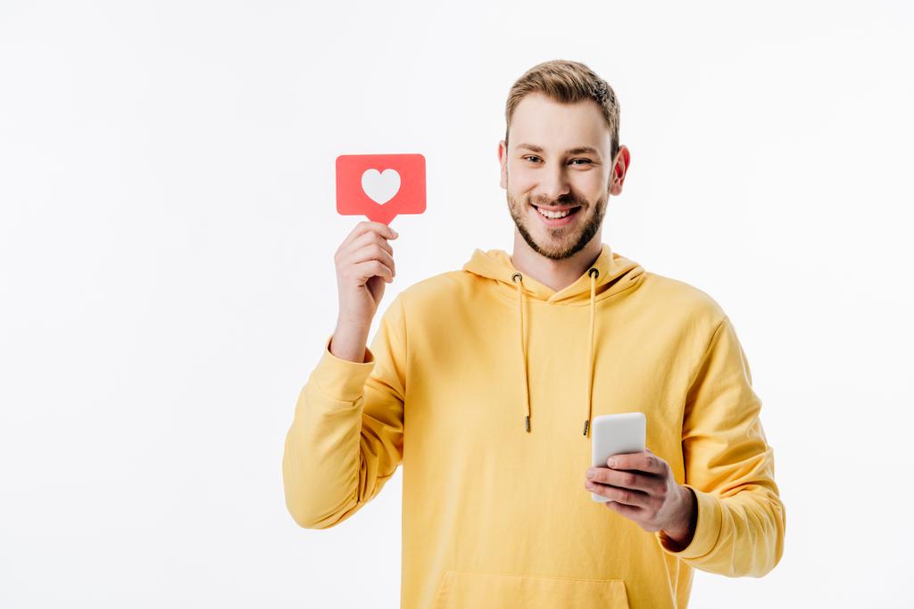 εύθυμος νεαρός άνδρας με κίτρινη κουκούλα κρατώντας κόκκινο χαρτί κομμένα κάρτα με το σύμβολο της καρδιάς, ενώ η χρήση smartphone απομονωθεί σε λευκό - Φωτογραφία, εικόνα