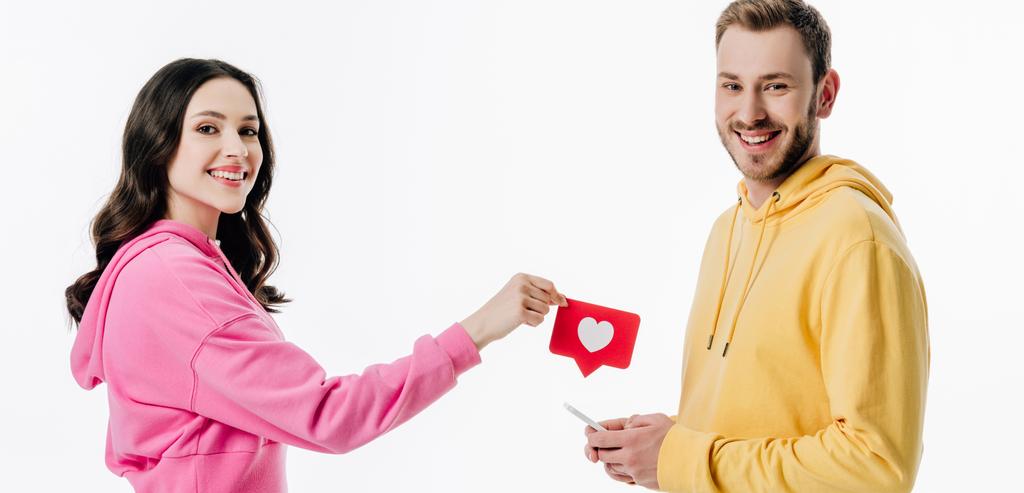 panoramiczny strzał uśmiechnięta dziewczyna dając czerwony papier cięcie karty z symbolem serca do przystojnego chłopaka z smartphone na białym tle - Zdjęcie, obraz