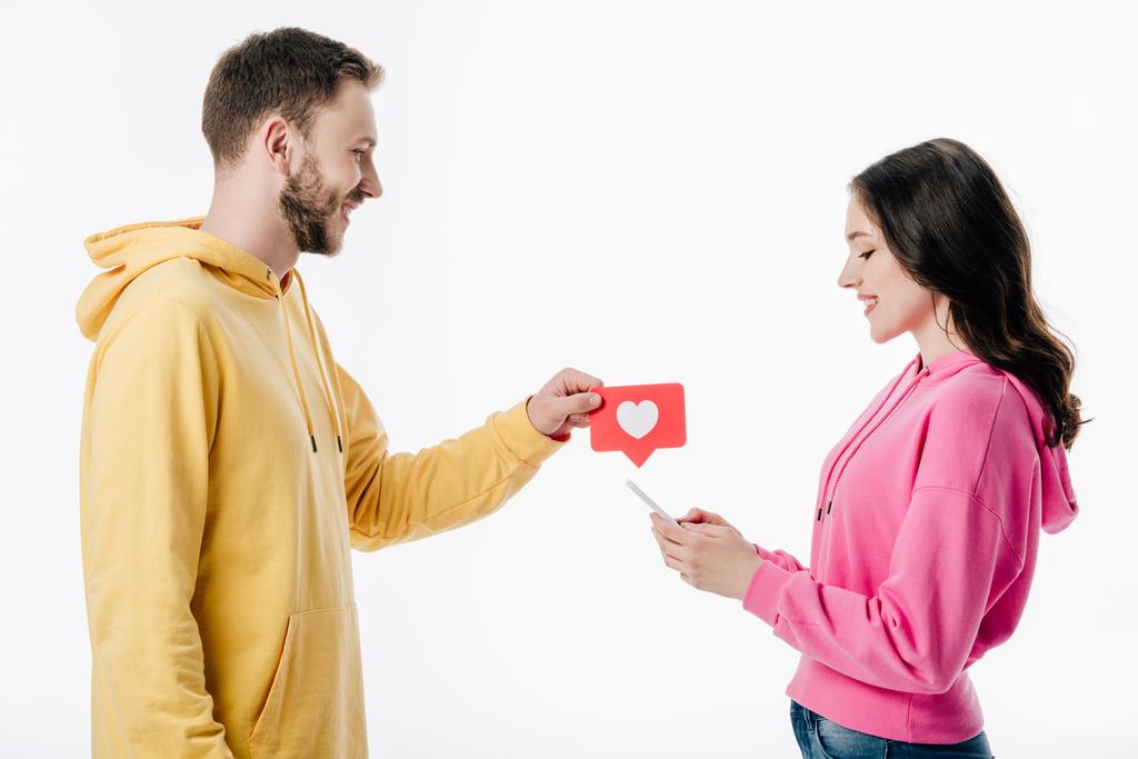 vista lateral de joven sonriente dando tarjeta de corte de papel rojo con símbolo del corazón a la novia usando teléfono inteligente aislado en blanco
 - Foto, imagen