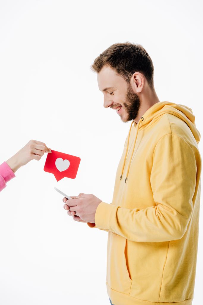 περικομμένη όψη της γυναίκας δίνοντας κόκκινο χαρτί κομμένα κάρτα με το σύμβολο της καρδιάς για τον νεαρό άντρα χρησιμοποιώντας smartphone απομονωθεί σε λευκό - Φωτογραφία, εικόνα