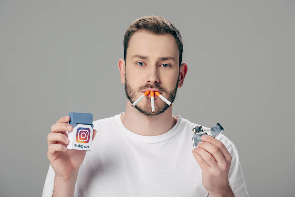 νεαρός άνδρας με τσιγάρα στο στόμα κρατώντας πακέτο με το λογότυπο Instagram και κοιτάζοντας κάμερα απομονωμένη σε γκρι - Φωτογραφία, εικόνα