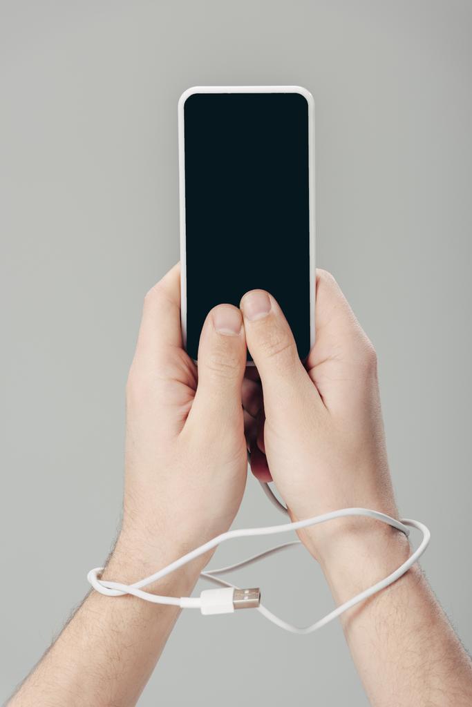 bijgesneden van de man met USB-kabel op handen houden smartphone met blanco scherm geïsoleerd op grijs - Foto, afbeelding