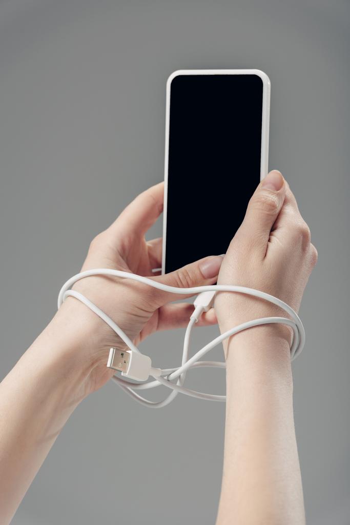 灰色で隔離された空白の画面を持つスマートフォンを持つ手の周りにUSBケーブルを持つ若い女性のトリミングビュー - 写真・画像