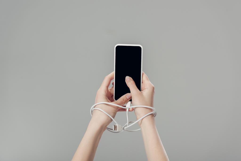 částečný pohled na mladou ženu s USB kabelem kolem rukou držící telefon Smartphone s prázdnou obrazovkou izolovanou na šedé - Fotografie, Obrázek
