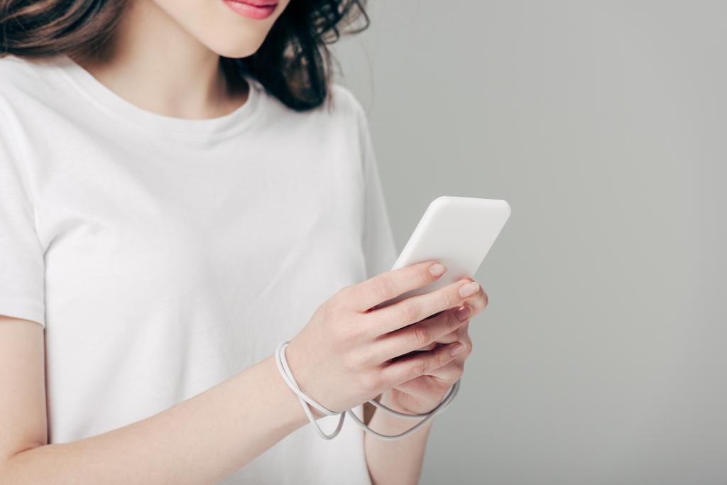 cropped view of young woman in white t-shirt and usb cable around hands using smartphone isolated on grey - Photo, Image