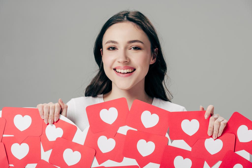 fröhliches junges Mädchen, das rote Karten mit Herzsymbol in der Hand hält und isoliert in die Kamera auf grau blickt - Foto, Bild