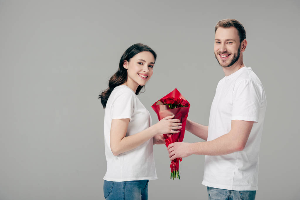 весёлые мужчина и женщина в белых футболках держат букет красных роз и смотрят на камеру изолированную на сером
 - Фото, изображение