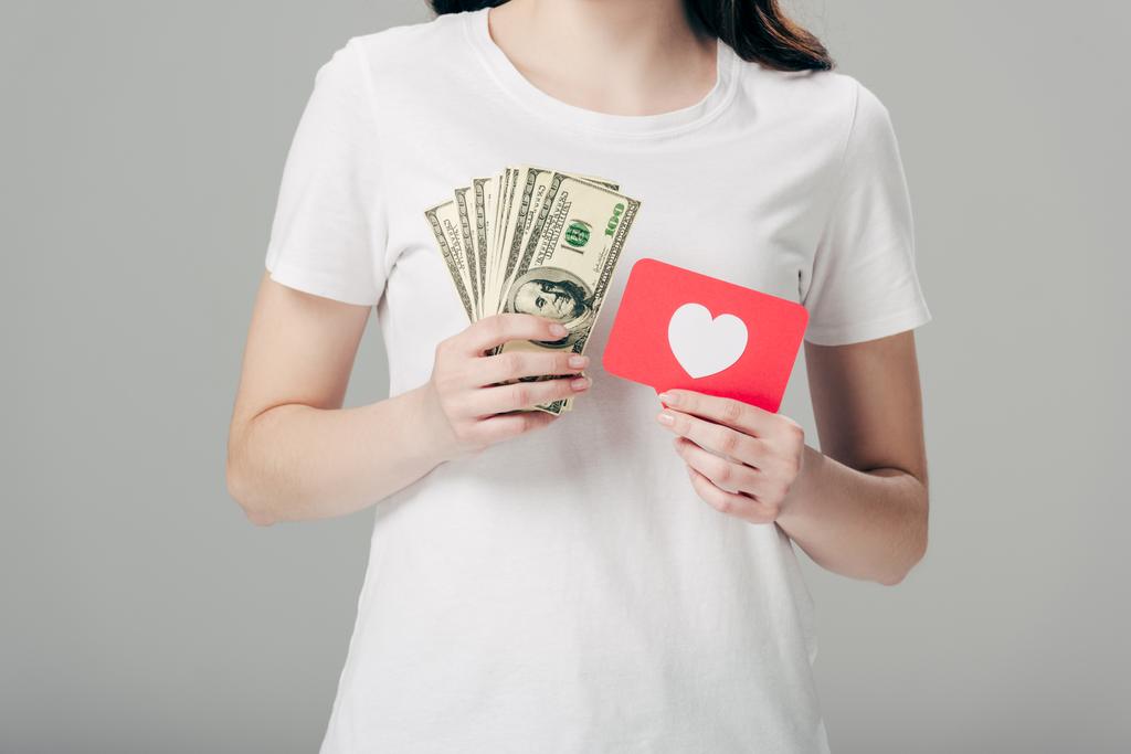 gedeeltelijke weergave van jonge meisje houden dollar bankbiljetten en rood papier knippen kaart met hart symbool geïsoleerd op grijs - Foto, afbeelding