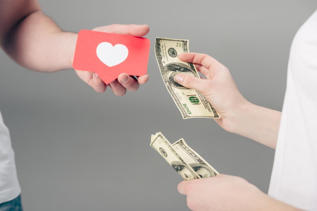 περικομμένη όψη της γυναίκας δίνοντας χαρτονομίσματα δολαρίων σε άνθρωπο με κόκκινο χαρτί κομμένο φύλλο με το σύμβολο της καρδιάς σε γκρίζο φόντο - Φωτογραφία, εικόνα