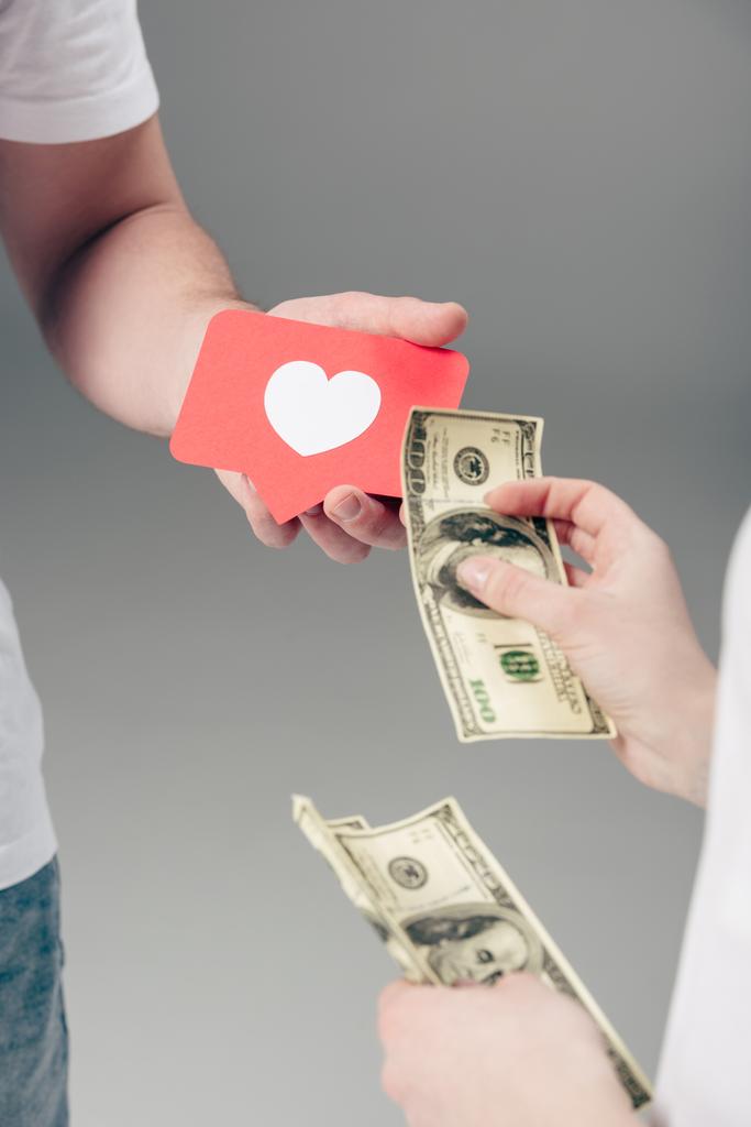 partial view of woman giving dollar banknotes to man with red paper cut card with heart symbol on grey background - Photo, Image