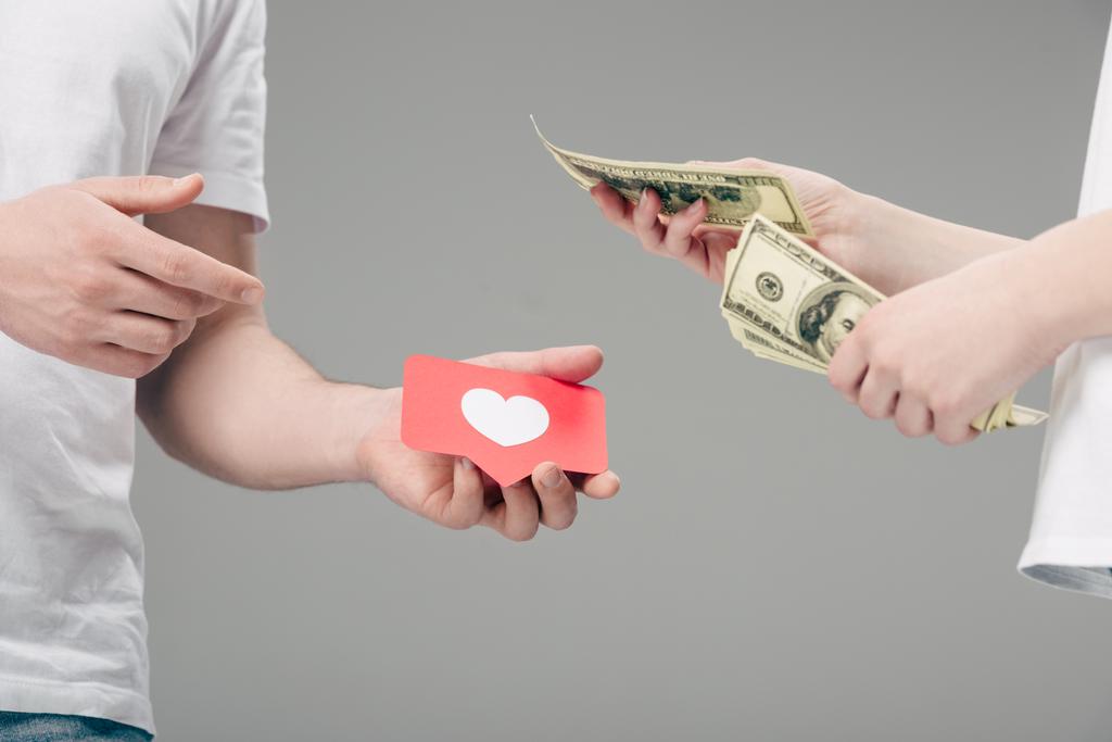 cropped view of woman giving dollar banknotes to man with red paper cut card with heart symbol isolated on grey - Photo, Image