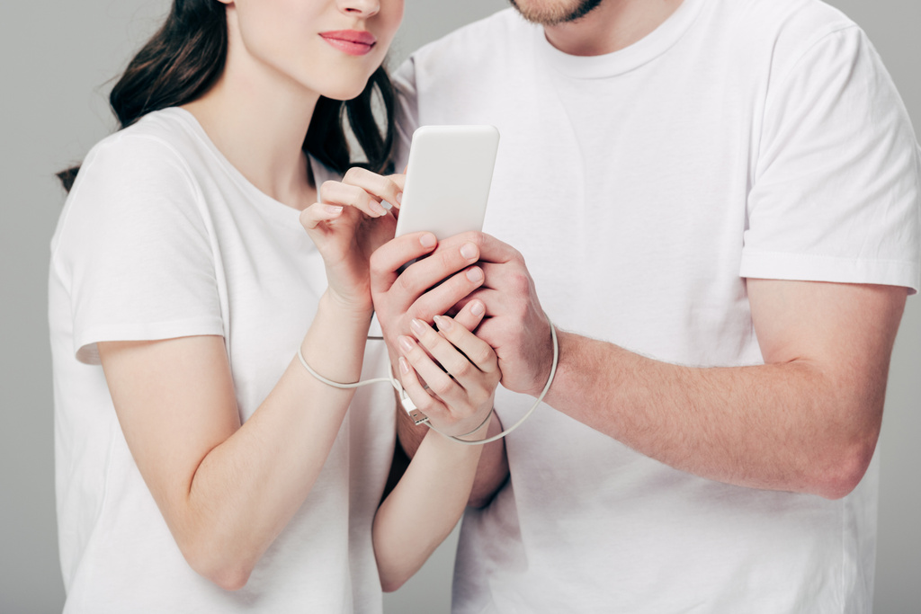 частичный вид мужчины и женщины с кабелем USB вокруг рук с помощью смартфона изолированы на сером
 - Фото, изображение
