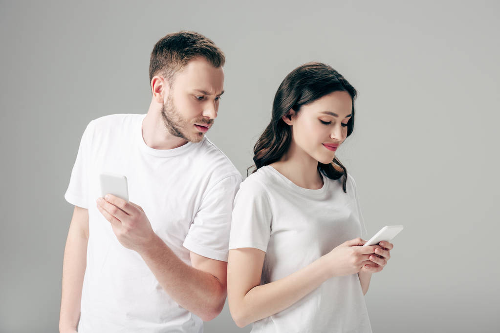 beledigd jonge man kijken naar mooie vriendin met behulp van smartphone geïsoleerd op grijs - Foto, afbeelding