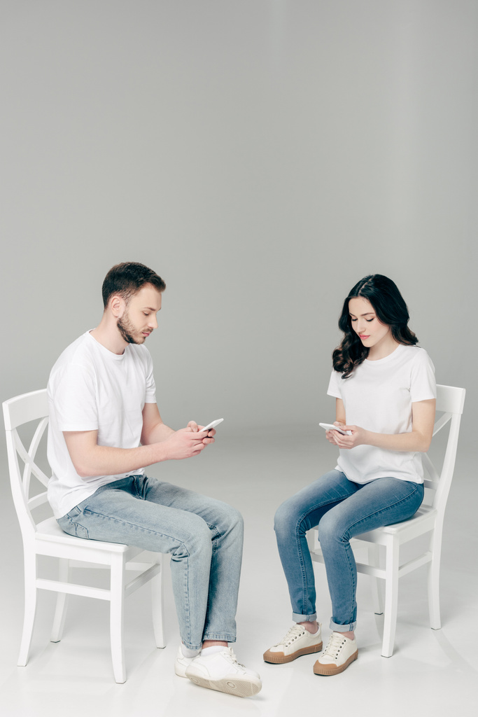 Junge Männer und Frauen in weißen T-Shirts und blauen Jeans sitzen auf Stühlen und nutzen Smartphones auf grauem Hintergrund. - Foto, Bild