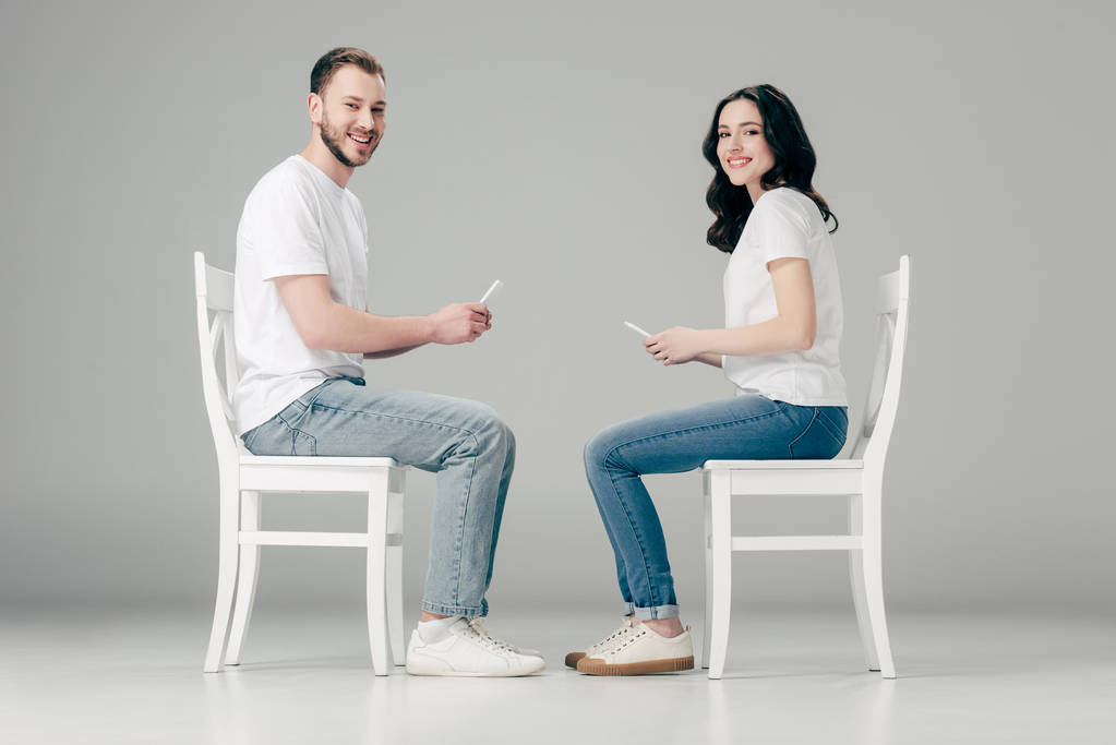 usmívající se muž a žena v bílých tričkách a modrých džínách, které sedí na židlích a používají telefony na šedé pozadí. - Fotografie, Obrázek