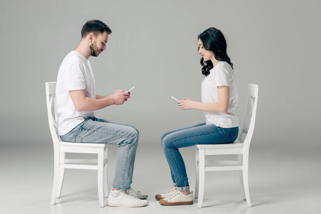 Zijaanzicht van geconcentreerde man en vrouw in witte t-shirts en blauwe jeans met behulp van smartphones, zittend op stoelen op grijze achtergrond  - Foto, afbeelding