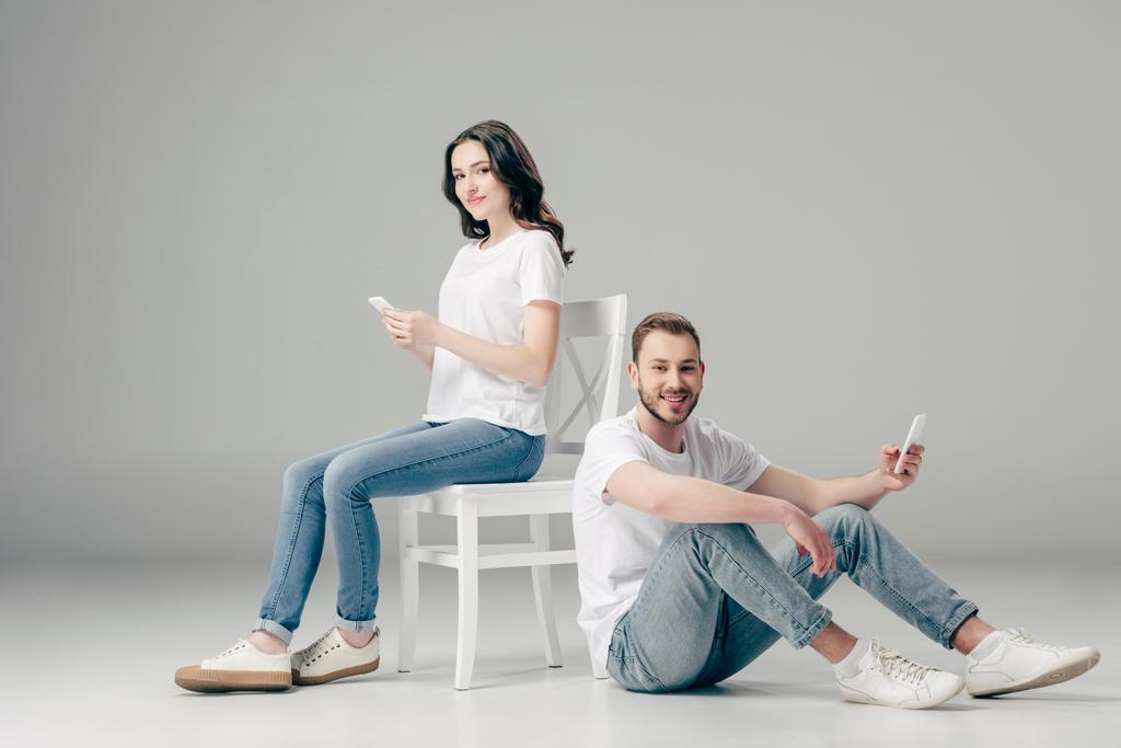 灰色の背景にスマートフォンで床に座っている陽気な男性の近くにスマートフォンを使用して笑顔の女性 - 写真・画像
