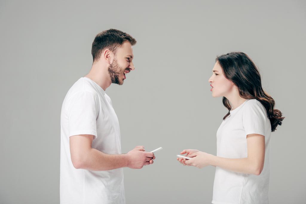vista lateral do jovem casal em camisetas brancas brigando enquanto segurando smartphones isolados em cinza
 - Foto, Imagem
