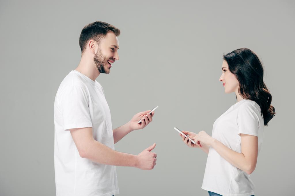hombre guapo mostrando el pulgar hacia arriba mientras está de pie cerca de chica bonita con teléfono inteligente aislado en gris
 - Foto, imagen