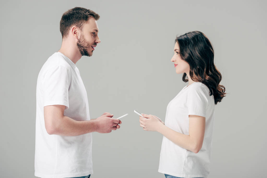 giovane uomo e donna in t-shirt bianche che si guardano mentre tengono gli smartphone isolati su grigio
 - Foto, immagini