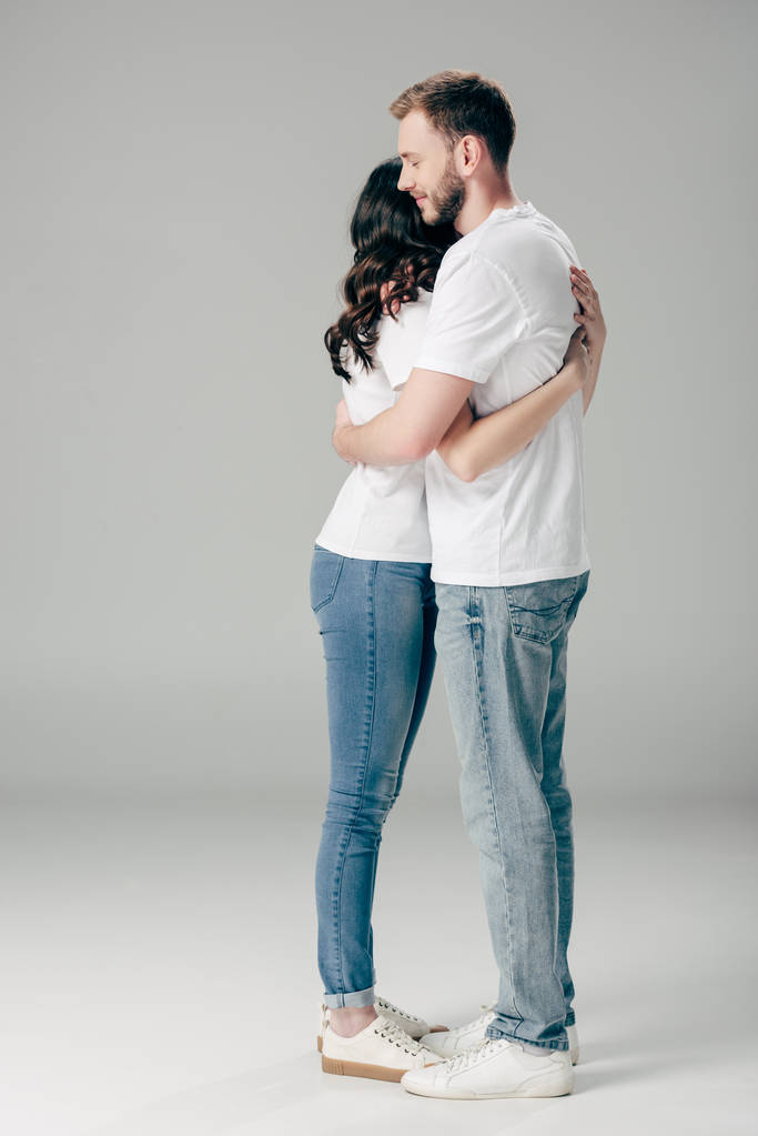 ευτυχισμένο νεαρό ζευγάρι με λευκά μπλουζάκια και μπλε τζιν που αγκαλιάζονται σε γκρίζο φόντο - Φωτογραφία, εικόνα