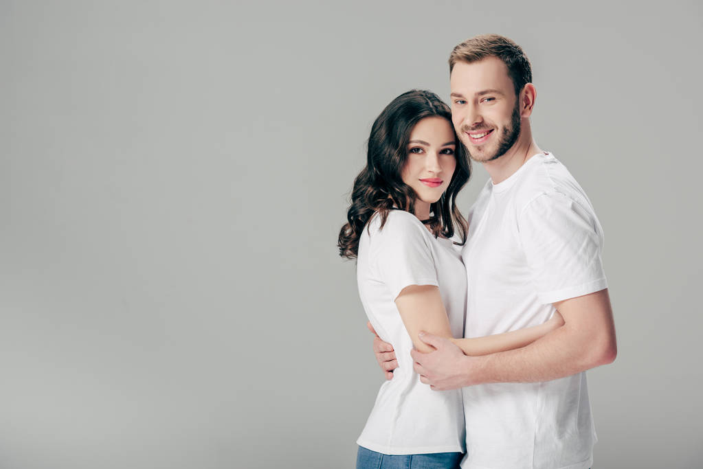 χαμογελαστή νεαρή ζευγάρι με άσπρα μπλουζάκια που αγκαλιάζονται και κοιτάζουν την κάμερα απομονωμένη σε γκρι - Φωτογραφία, εικόνα