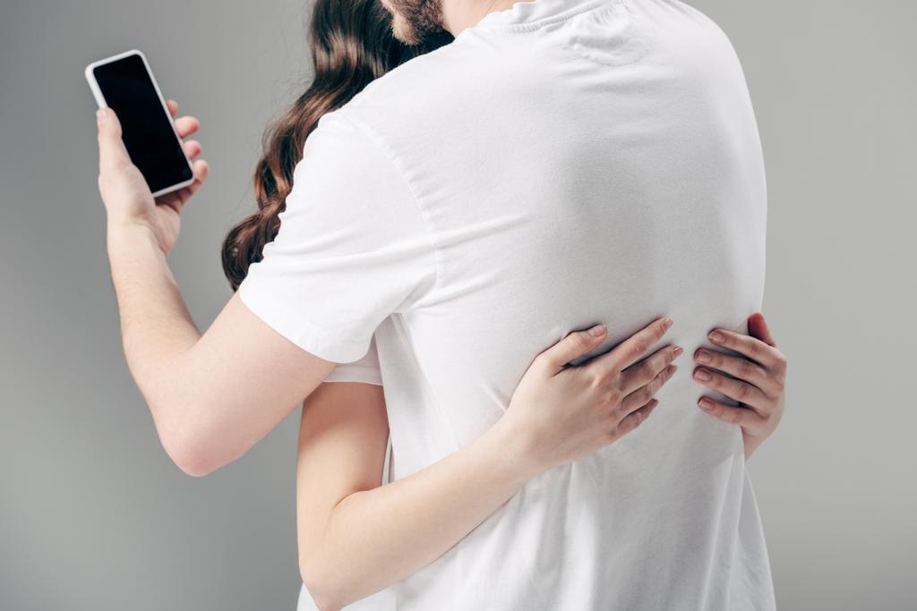 vista recortada de la mujer abrazando al hombre usando un teléfono inteligente sobre fondo gris
 - Foto, Imagen