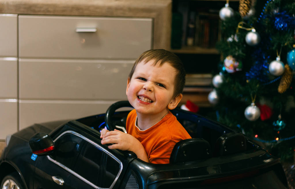 Un bambino è seduto in una macchinina, vicino all'albero di Natale. Un regalo per il nuovo anno. la smorfia gioisce
 - Foto, immagini