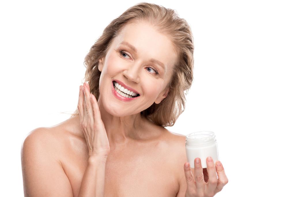 όμορφη ώριμη γυναίκα με τέλειο δέρμα εφαρμόζοντας καλλυντική κρέμα και χαμογελαστά απομονωμένα σε λευκό - Φωτογραφία, εικόνα