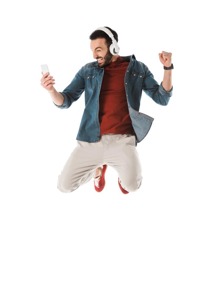 excited man in headphones jumping while using smartphone and showing yes gesture isolated on white - Photo, Image