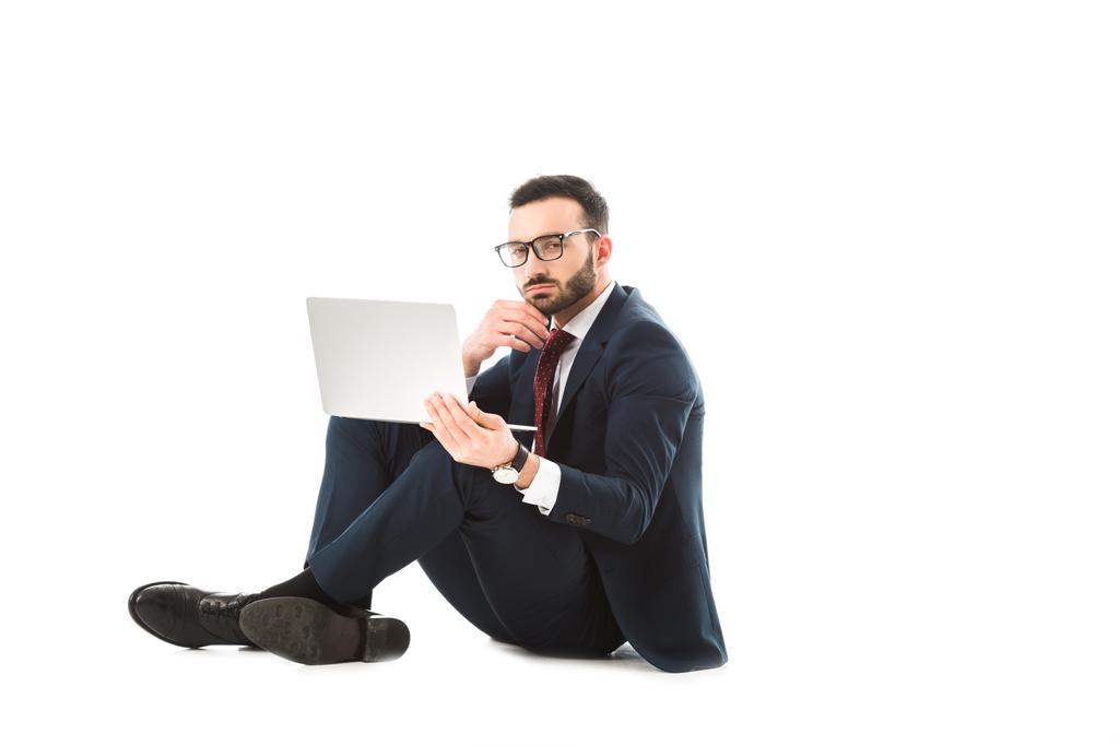 doordachte zakenman met behulp van laptop en kijken naar camera terwijl zittend op witte achtergrond - Foto, afbeelding
