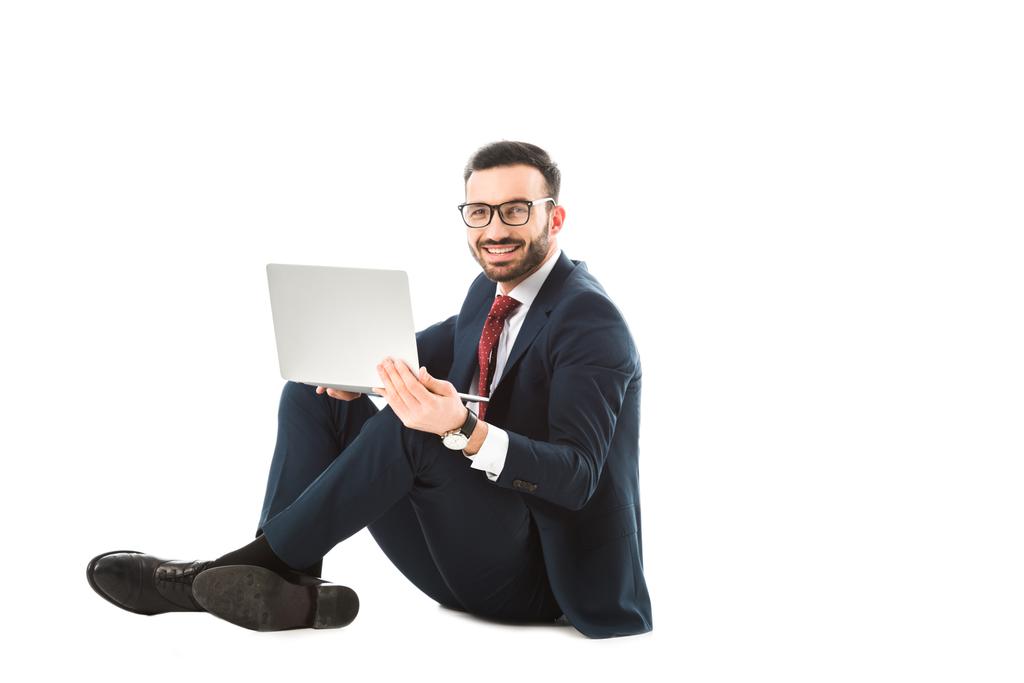 lachende zakenman kijkt naar camera tijdens het zitten en het gebruik van laptop op witte achtergrond - Foto, afbeelding
